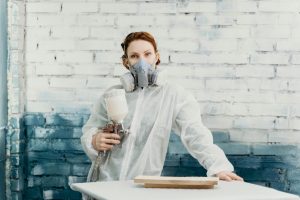 Woman in protective suit and gas mask using spray gun indoors against a painted brick wall. Can you Spray Laminate Kitchen Cabinet